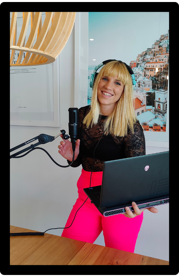 A smiling woman with blonde hair, wearing a black lace top and bright pink pants, stands in a well-lit room using a microphone and holding a laptop, displaying a coming soon page.